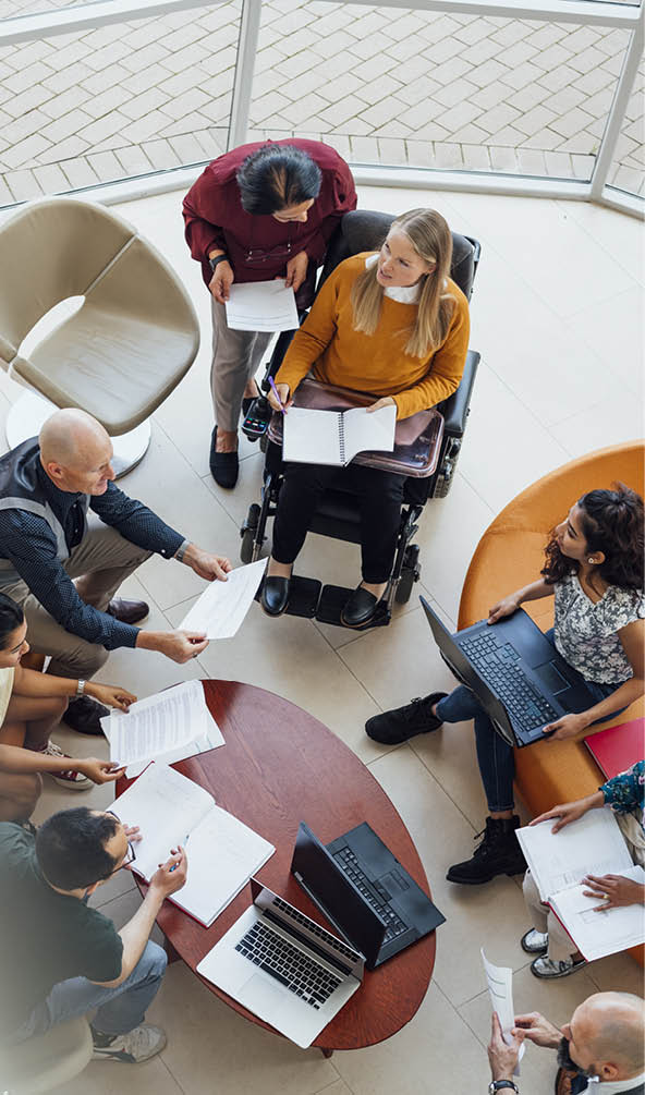 A professional team hold their regular meeting  They are a diverse group of multi-ethnicities and age ranges  They can be seen mid-discussion and are holding documents and laptops 