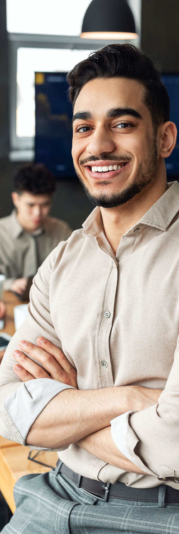 Successful Person  Portrait of confident smiling bearded businessman sitting leaning on desk in office, posing with folded arms and looking at camera, colleagues working in blurred background