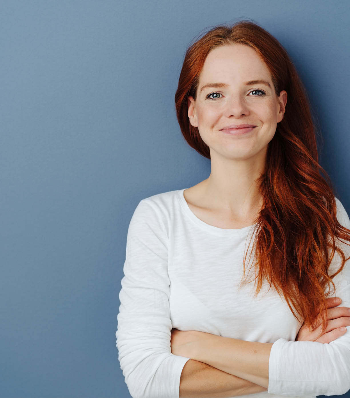 Pleased confident young redhead woman with a beaming smile and folded arms posing on a blue studio background with copy space grinning at the camera